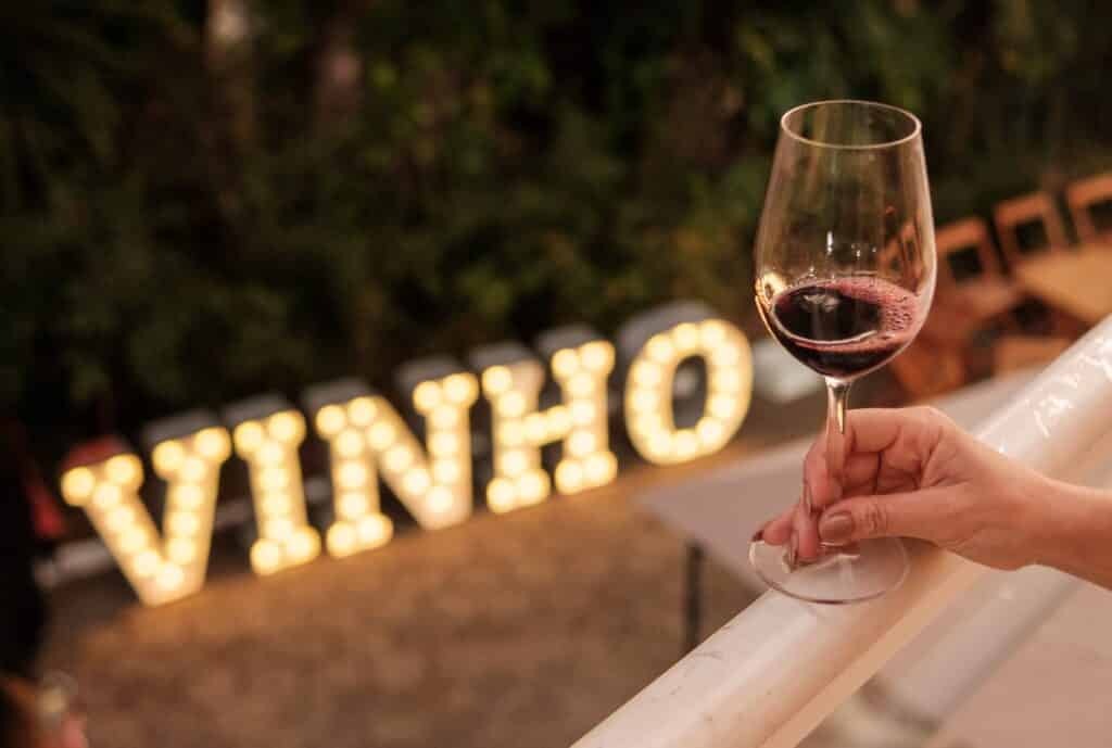Taça de vinho tinto sendo servida no evento Vinho na Vila, com letreiro luminoso ao fundo