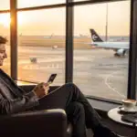 Viajante lendo notícia no smartphone dentro de sala VIP de aeroporto premium, com cartão preto sobre a mesinha e vista para a pista.